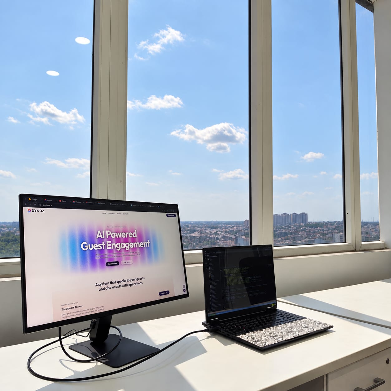 A monitor showing the Dynoz site and an open laptop on a desk at the window, under open sky.